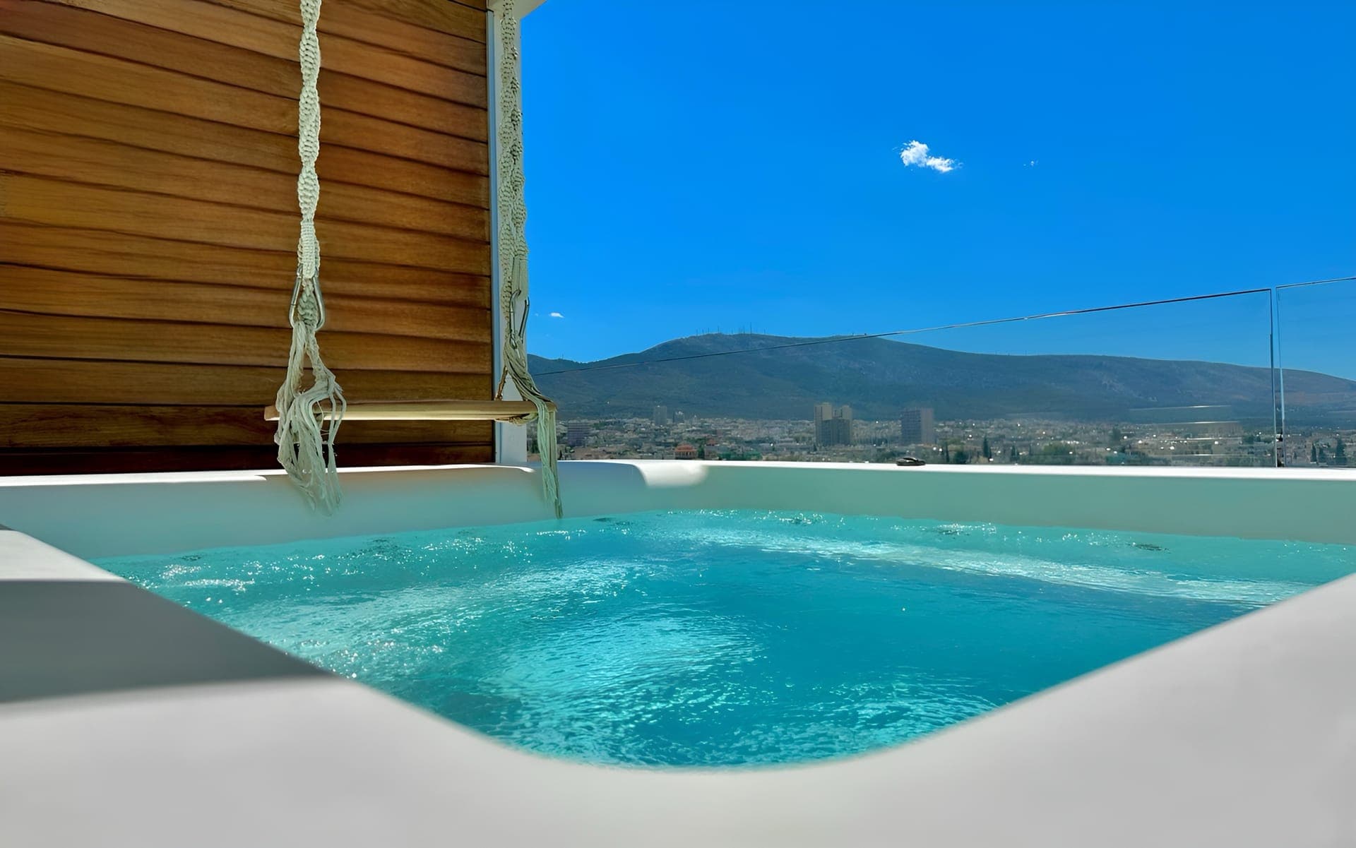 Our serene outdoor spacious all season jacuzzi with crystal-clear water, surrounded by a wooden deck and a hanging chair, set against a backdrop of mountains under a bright blue sky