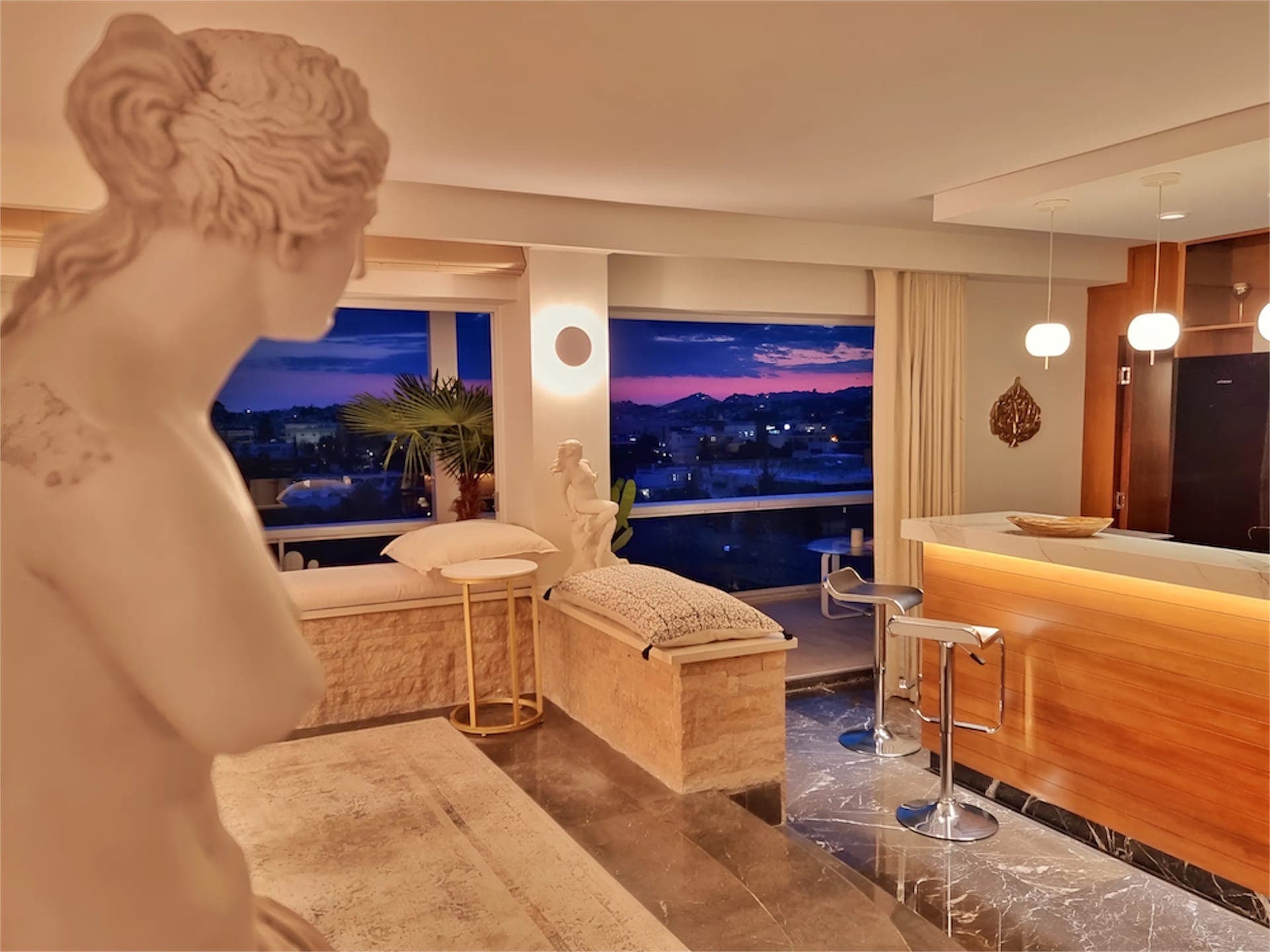 Our luxurious indoor living space featuring elegant décor, including a classical statue, plush seating, and large windows offering a stunning view of the cityscape at dusk