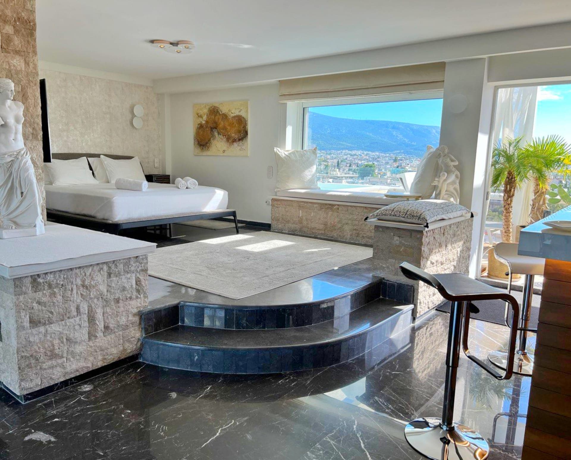 Our cozy open plan living space featuring a king sized bed, classical statues, a flat-screen TV, a bar, with the view of a cityscape and mountains in the background